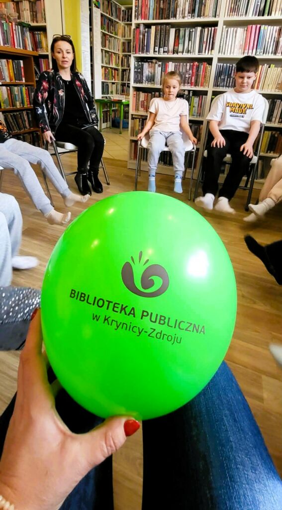 Dzieci siedza w sali bibliotecznej. na krzesłacj. jedna osoba trzyma w ręce balon zielony z logiem biblioteki w Krynicy Zdroju