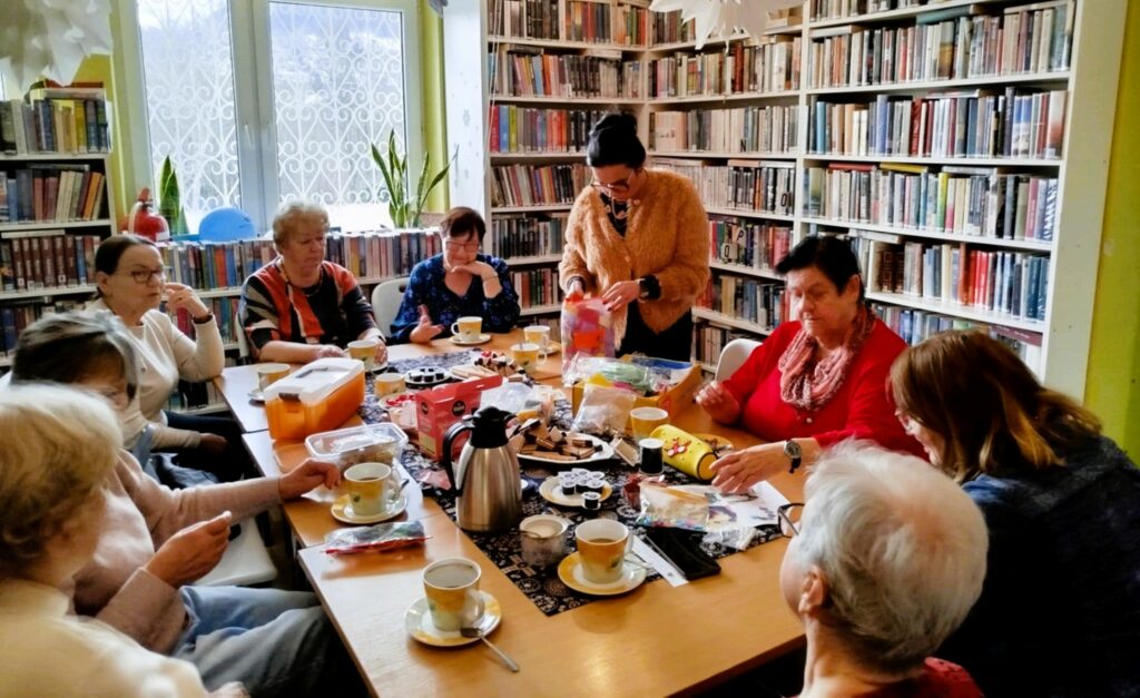 na zdjęciu widać panie seniorki siedzące wokół stołu w sali bibliotecznej i wykonujące korale
