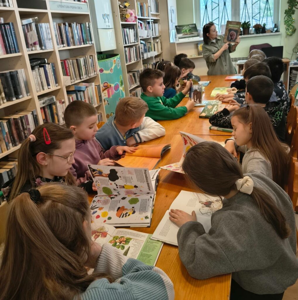 Przy długim stole siedzą dzieci w wieku szkolnym. oglądają ksiązki . Pani bibliotekarka prezentuję książkę