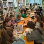 Przy długim stole siedzą dzieci w wieku szkolnym. oglądają ksiązki . Pani bibliotekarka prezentuję książkę