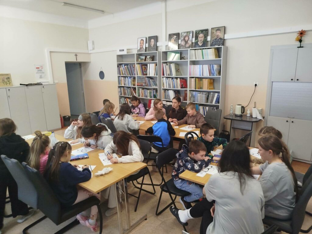 Duża grupa dzieci siedzi przy stołach i wykonuje prace plastyczne . za nimi regał z książkami.