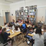 Duża grupa dzieci siedzi przy stołach i wykonuje prace plastyczne . za nimi regał z książkami.