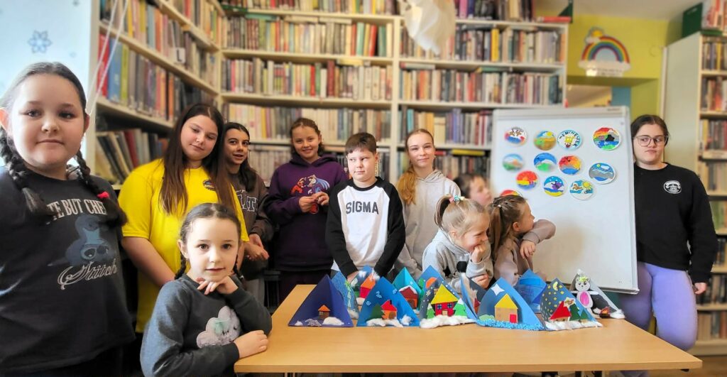 na zdjęciu widać dzieci w sali bibliotecznej stojących na tle regału z książkami