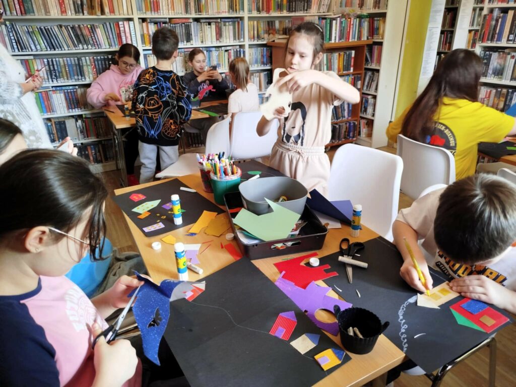 Przy stołach w sali bibliotecznej siedzą dzieci i wykonuja prace plastyczne