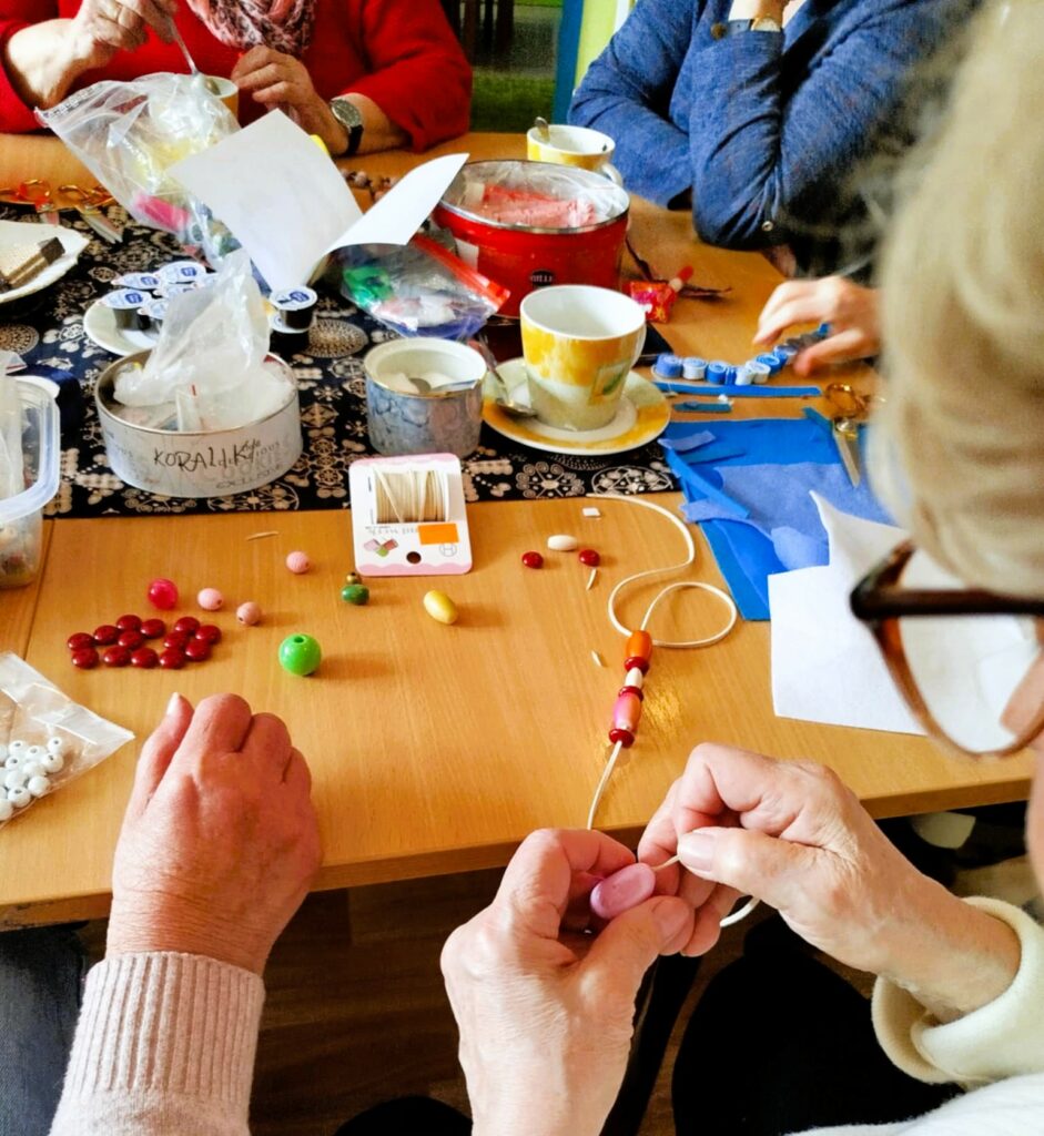 na zdjęciu widać panie seniorki siedzące wokół stołu w sali bibliotecznej i wykonujące korale