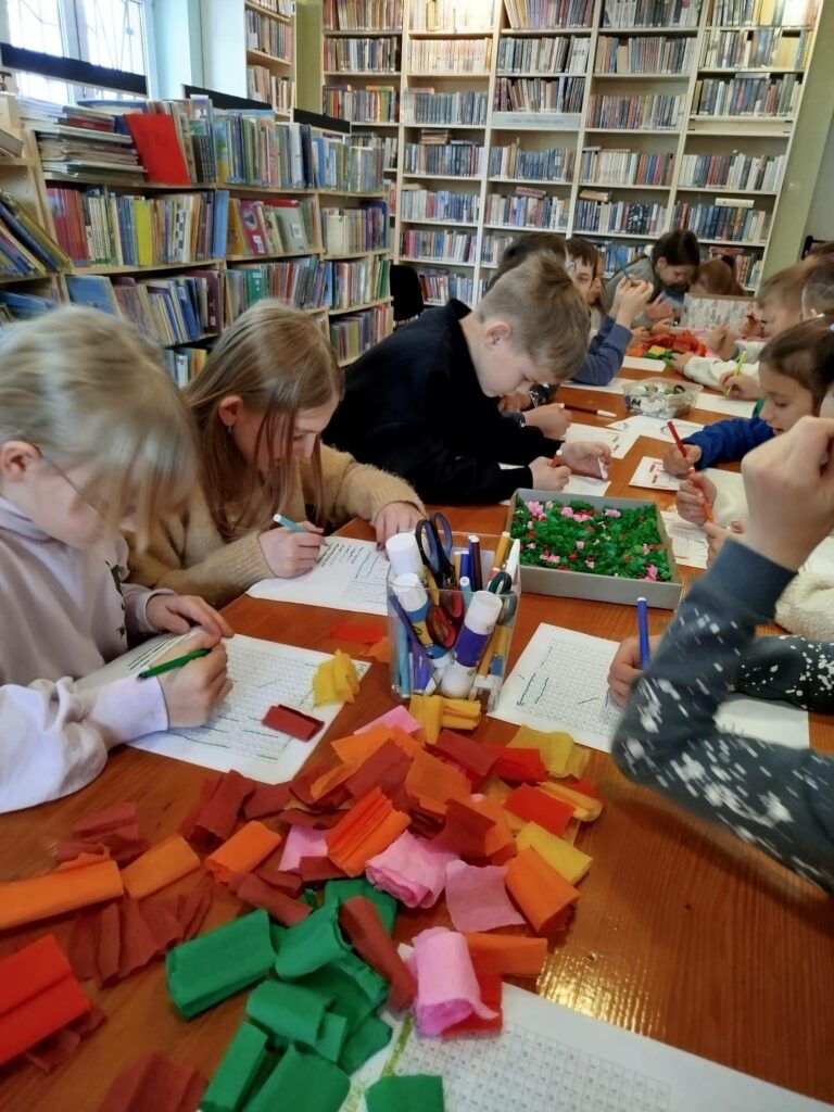 Przy stołach w sali bibliotecznej siedza dzieci w wieku szolnym. Dzieci rozwiązują rebusy. na stole bibułai akcesoria do malowania.