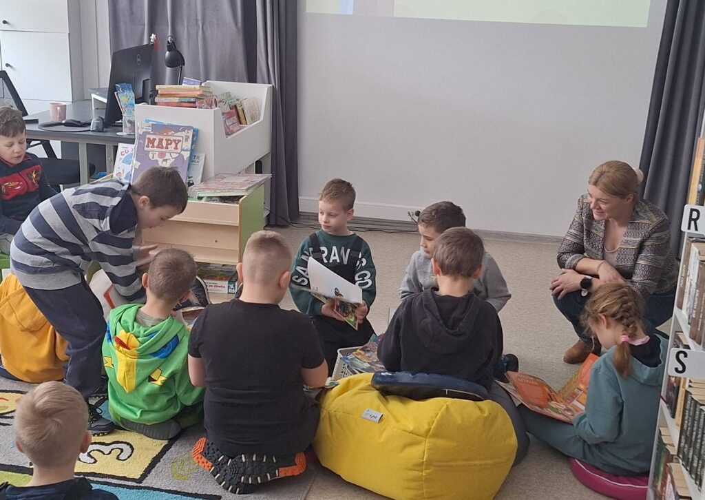 Na ziemi i pufach siedzi grupa dzieci wraz z panią bibliotekarka. Wszyscy oglądaja kolorowe bajki.