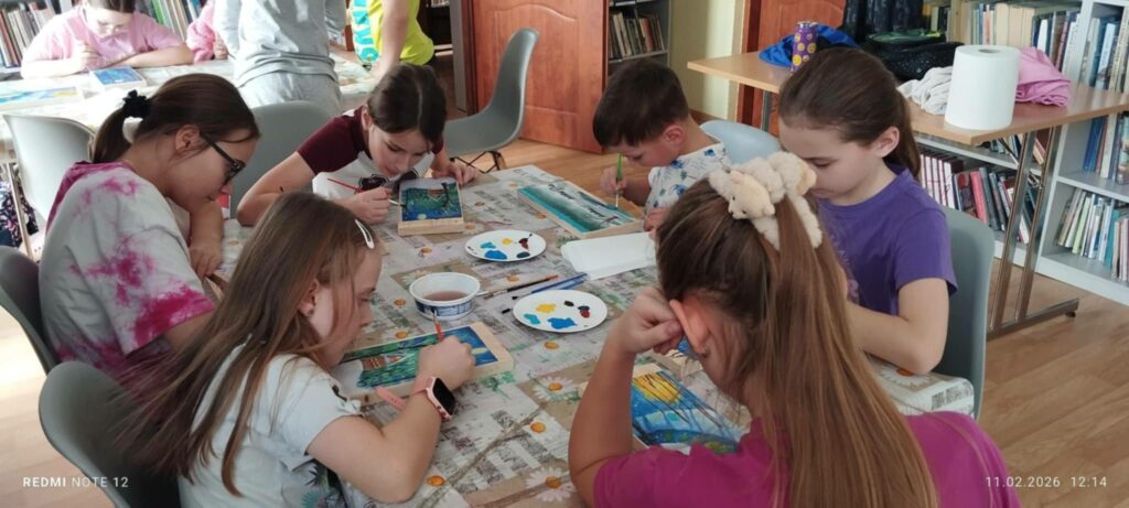 Przy stołach w sali bibliotecznej dzieci siedza przy stołach i wykonują prace plastyczne.