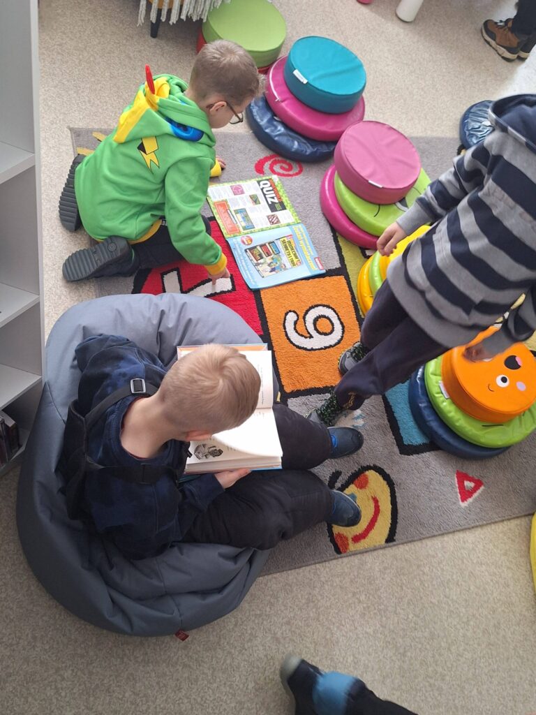 na zdjęciu dzieci siedzące na ziemi w sali bibliotecznej. dwóch chłopców czyta ksiązki jeden stoi . Wokół kolorowe pufy do siedzenia