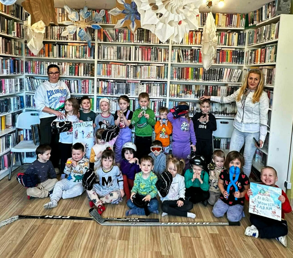 na zdjęci na tle regałów z książkami siedza i stoją dzieci wraz z dwiema paniami do wspólnego zdjęcia.