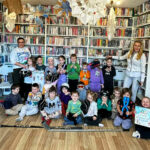 na zdjęci na tle regałów z książkami siedza i stoją dzieci wraz z dwiema paniami do wspólnego zdjęcia.