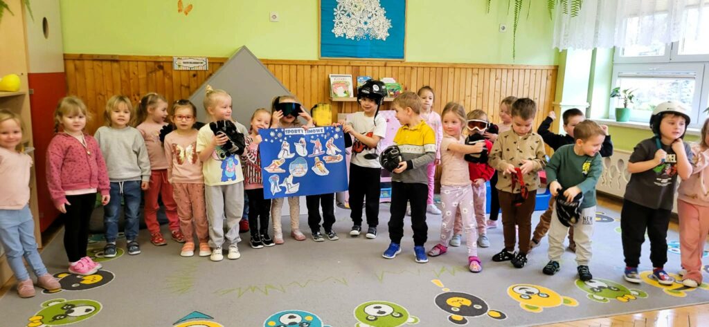 na zdjęciu grupa dzieci w wieku przedszkolnym ustawiona do wspólnego zdjęcia. Niektóre trzymaja w ręku akcesoria sportowe miedzy innymi kask na głowe , gogle , rękawice