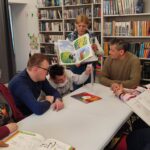 Przy stole siedzi trzech mężczyzn. Pani stoi przy nich i pokazuje im ksiązkę. W tyle regały z ksiązkami.