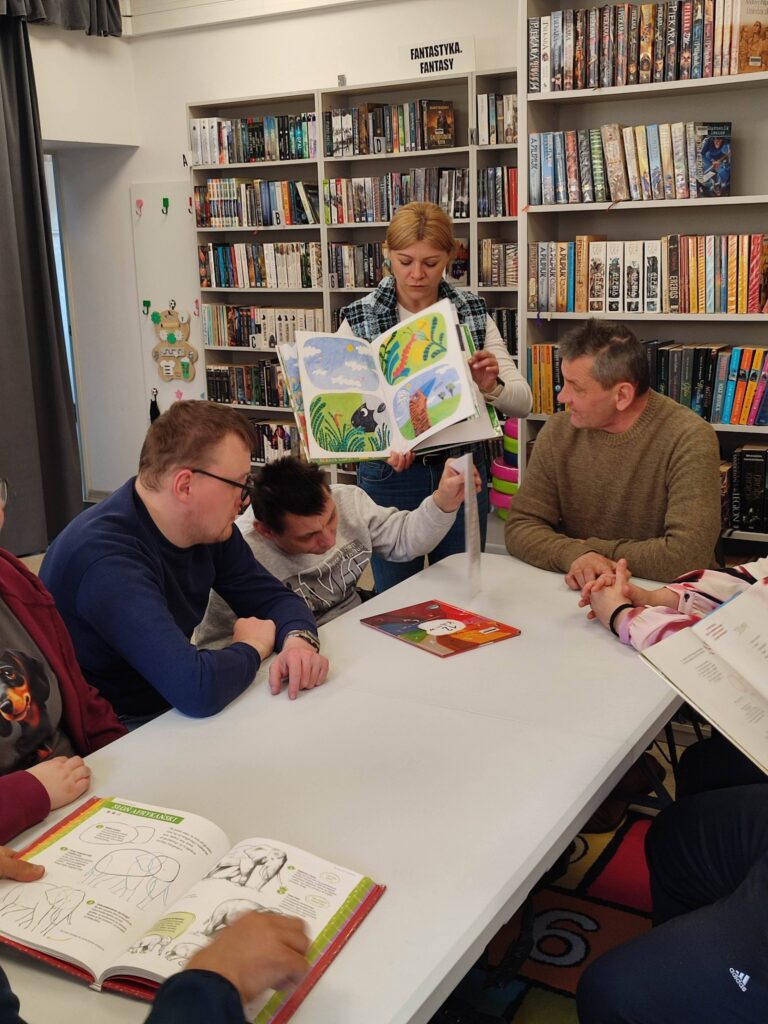 Przy stole siedzi trzech mężczyzn. Pani stoi przy nich i pokazuje im ksiązkę. W tyle regały z ksiązkami.