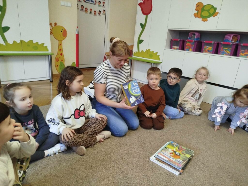 Na ziemi wraz z dziećmi w kółeczku siedzi pani bibliotekarka i pokazuje dzieciom książeczkę o kotach