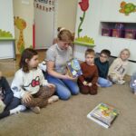 Na ziemi wraz z dziećmi w kółeczku siedzi pani bibliotekarka i pokazuje dzieciom książeczkę o kotach