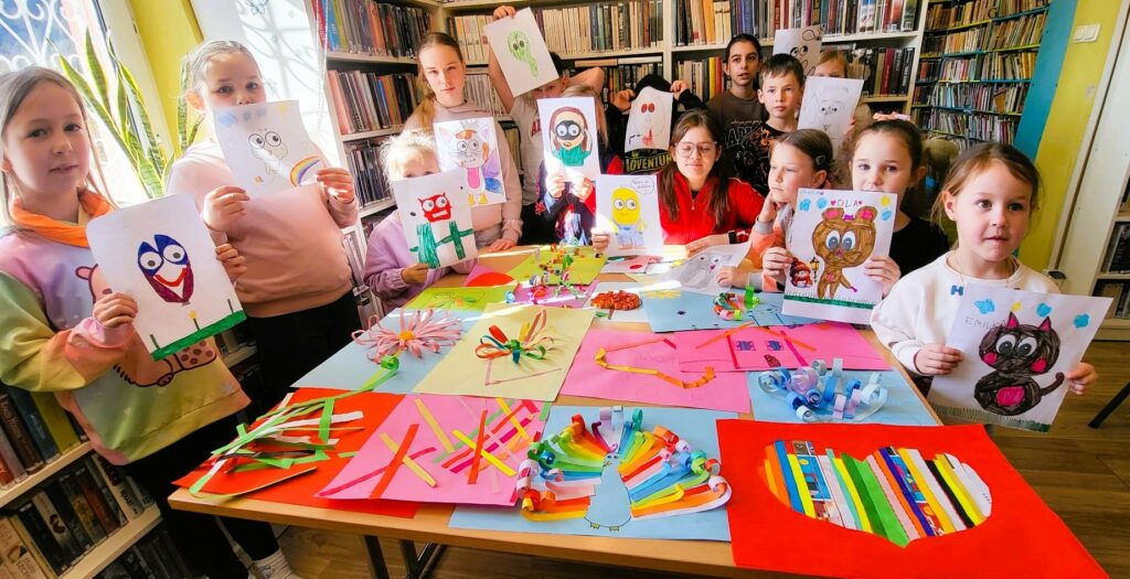 Dzieci przedszkolne w sali bibliotecznej prezentują wykonane przez siebie prace - wyklejankina kolorowych papierach.