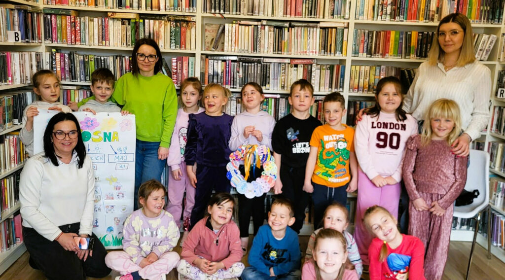 Na tle regałów stoją dzieci wraz z paniami bibliotekarkami i wychowawczynią