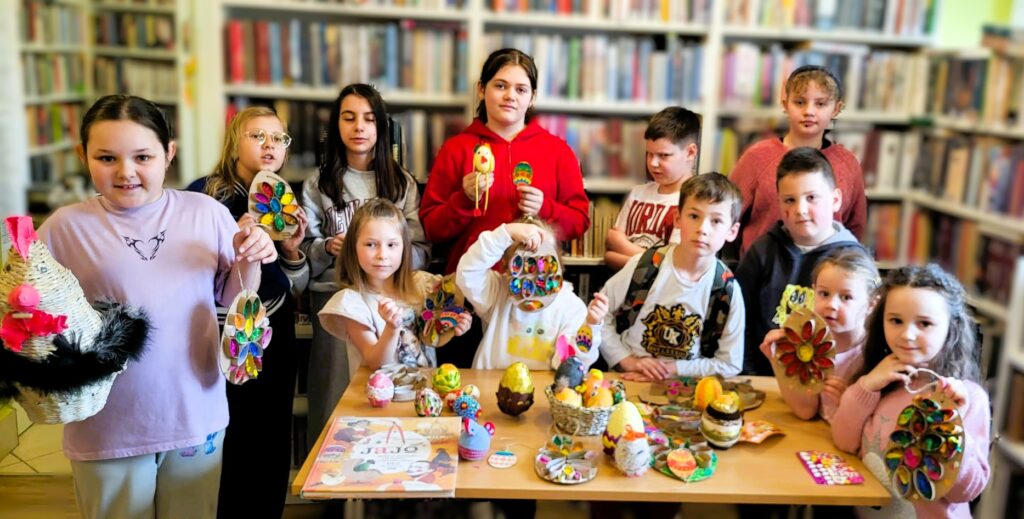 na zdjęciu widać stół na którym poukładano jajeczka wykonane różnymi metodami plastycznymi. W tyle stoją dzieci uczestniczące w zajęciach, prezentują wykonane stroiki świąteczne