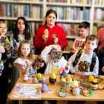 na zdjęciu widać stół na którym poukładano jajeczka wykonane różnymi metodami plastycznymi. W tyle stoją dzieci uczestniczące w zajęciach, prezentują wykonane stroiki świąteczne