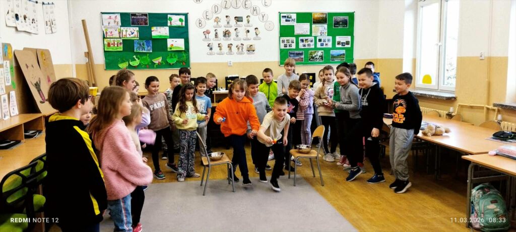 W sali w szkole duża grupa dzieci stoi i po kolei biara udział w rywalizacji w róznych konkurencjach. Na środku dwa krzesła na , których sa miseczki. Za nimi na ścianach gazetki z kolorowymi ilustracjami.