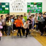 W sali w szkole duża grupa dzieci stoi i po kolei biara udział w rywalizacji w róznych konkurencjach. Na środku dwa krzesła na , których sa miseczki. Za nimi na ścianach gazetki z kolorowymi ilustracjami.