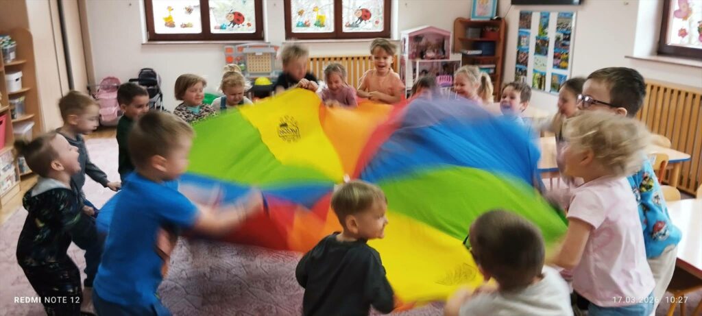 W sali dzieci stoja w kółku. W środku jest kolorowa chusta, która za końce trzymaja dzieci zgromadzone wokół.