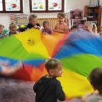 W sali dzieci stoja w kółku. W środku jest kolorowa chusta, która za końce trzymaja dzieci zgromadzone wokół.