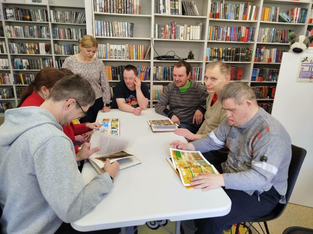 Przy stole siedzą młode osoby . Cztery osoby oglądają ksiązki kolorowe. W tyle regały z kolorowymi książkami