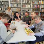 Przy stole siedzą młode osoby . Cztery osoby oglądają ksiązki kolorowe. W tyle regały z kolorowymi książkami