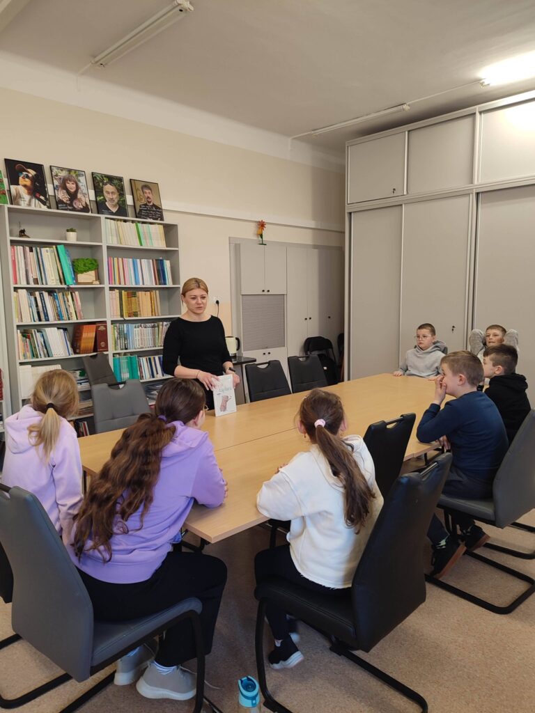 Przy stole siedmioro dzieci . Przed nimi stoi pani i prezentuje ksiązkę. Za nią regały z książkami