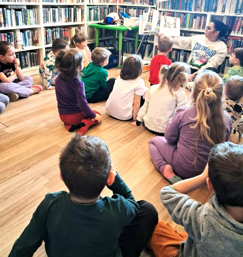 W sali bibliotecznej na ziemi siedzi pani bibliotekarka i czyta dzieciom książke. Wokół niej dzieci słuchające opowieści.