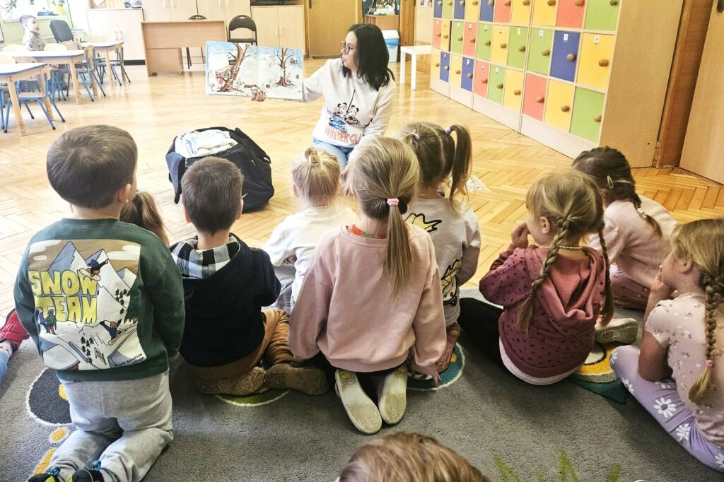 Na zdjęciu widać bibliotekarke czytającą książkę oraz dzieci siedzące i słuchające opowieści.
