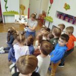 Stoi wraz z dziećmi w kółeczku pani bibliotekarka i pokazuje dzieciom książeczkę o kotach Wokół kolorowa sala.