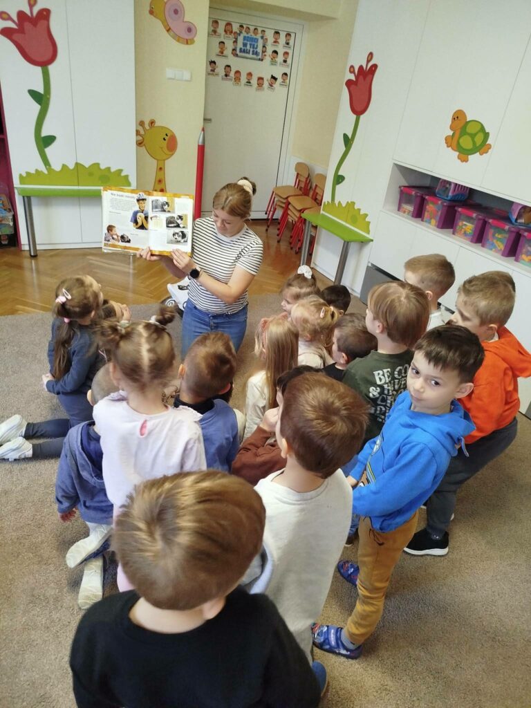 Stoi wraz z dziećmi w kółeczku pani bibliotekarka i pokazuje dzieciom książeczkę o kotach Wokół kolorowa sala.
