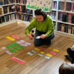 Na zdjęciu pani bibliotekarka pochylona nad kolorowymi kartkami . W tyle za nią prezentowany plakat z wiosną i regały z kolorowymi książkami