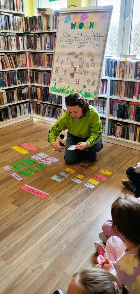 Na zdjęciu pani bibliotekarka pochylona nad kolorowymi kartkami . W tyle za nią prezentowany plakat z wiosną i regały z kolorowymi książkami