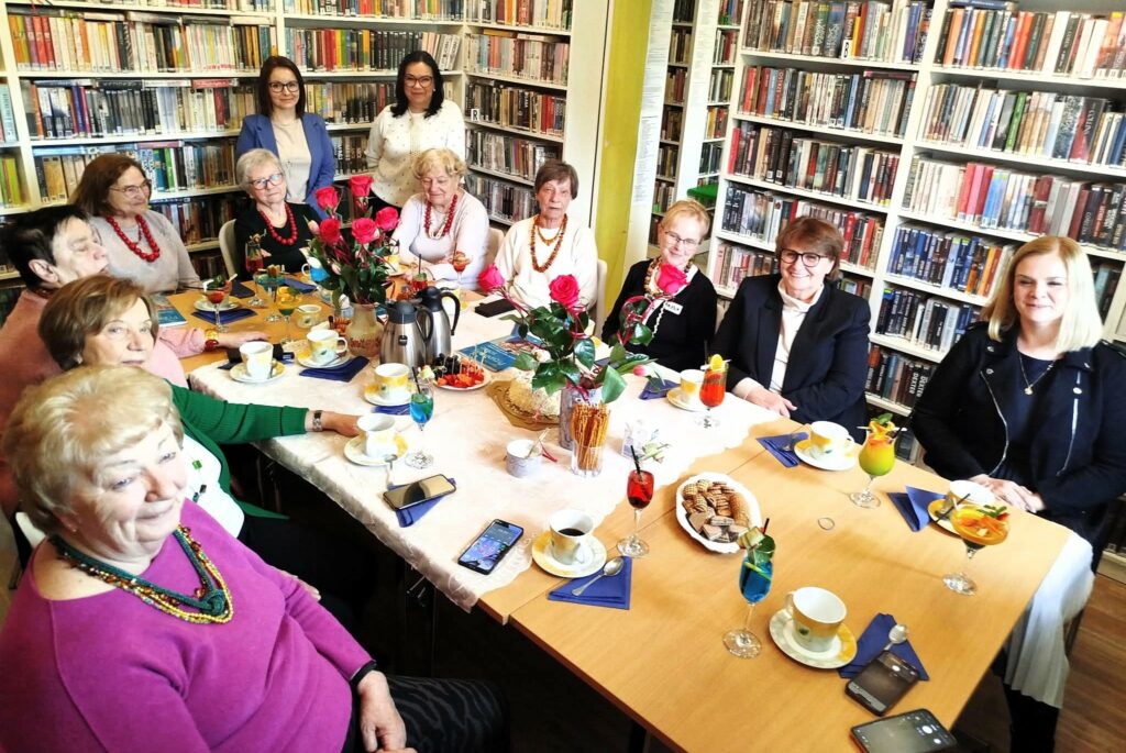 na zdjęciu widać grono kobiet siedzące wokół stołu w sali bibliotecznej. Na sole kawa w filiżankach, kwiaty, ciastka. Wokół regały z kolorowymi książkami.i
