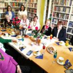 na zdjęciu widać grono kobiet siedzące wokół stołu w sali bibliotecznej. Na sole kawa w filiżankach, kwiaty, ciastka. Wokół regały z kolorowymi książkami.i