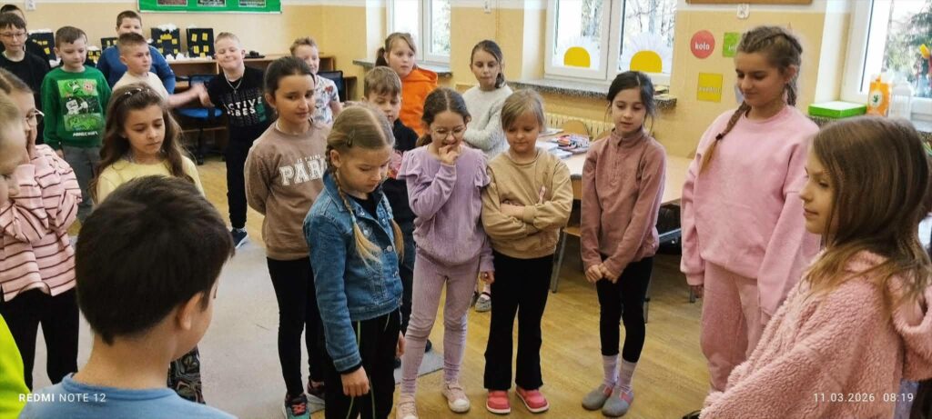 W sali stoi grupa dzieci w kółku . Wszyscy ubrani w kolorowe bluzy . Wzrok mają skierowany na ziemię.