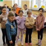 W sali stoi grupa dzieci w kółku . Wszyscy ubrani w kolorowe bluzy . Wzrok mają skierowany na ziemię.