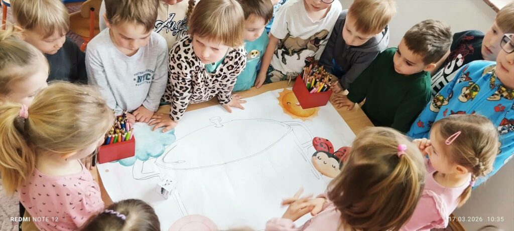 Na stole jest duży karton na nim ilustracja do pokolorowania z boku kredki . Wokoł stołu dzieci przyglądające się rysunkowi