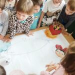 Na stole jest duży karton na nim ilustracja do pokolorowania z boku kredki . Wokoł stołu dzieci przyglądające się rysunkowi