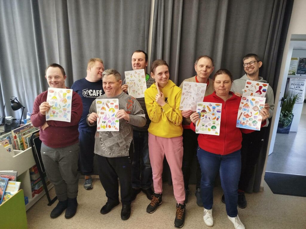 osiem osób stoi na tle szarej zasłony i prezentują prace plastyczne.