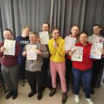 osiem osób stoi na tle szarej zasłony i prezentują prace plastyczne.