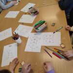 Na stole porozkładane karty z zabawali logicznymi. widać flamastry, kredki oraz dzieciece ręce , które wykonuja zagadki.