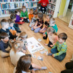 Drupa przedszkolaków siedzi na podłodze w sali bibliotecznej na ziemi w kółeczku i gra w grę. Za nimi regał z książkami .