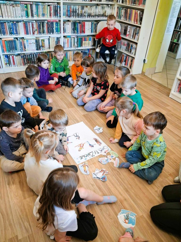Drupa przedszkolaków siedzi na podłodze w sali bibliotecznej na ziemi w kółeczku i gra w grę. Za nimi regał z książkami .