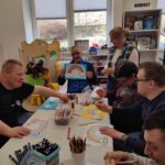 Przy stole wokół siedza młodzi ludzie i wykonuja prace plastyczną tęczę z chmurkami. Na stole flamastry i kredki