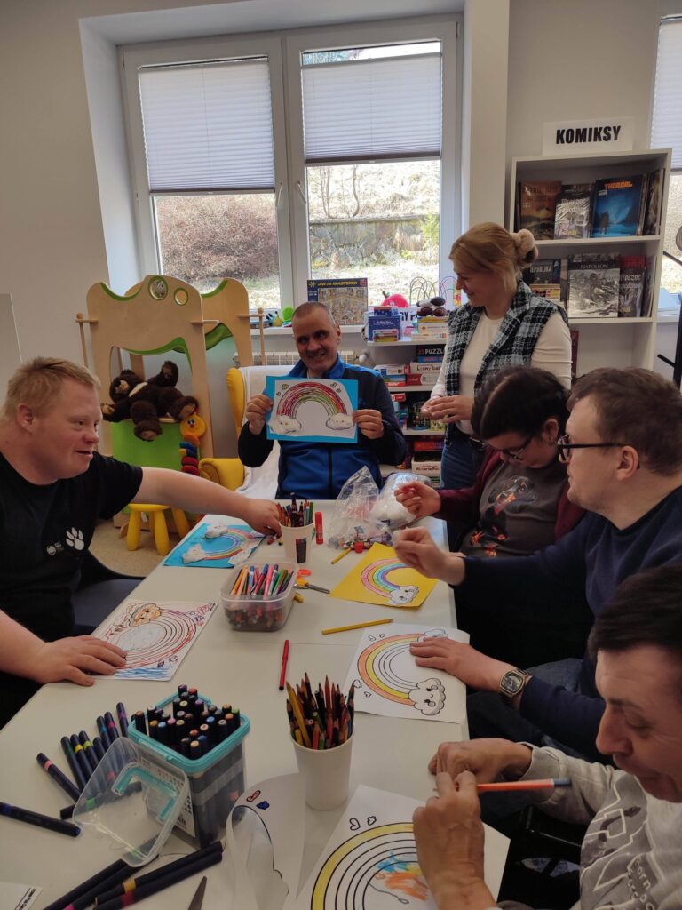 Przy stole wokół siedza młodzi ludzie i wykonuja prace plastyczną tęczę z chmurkami. Na stole flamastry i kredki